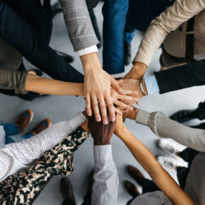A high-angle shot of a group of male and female colleagues putting their hands together in an office. They are dressed in fashionable business clothes. Their faces are not visible, only their arms. Horizontal daylight indoor photo.